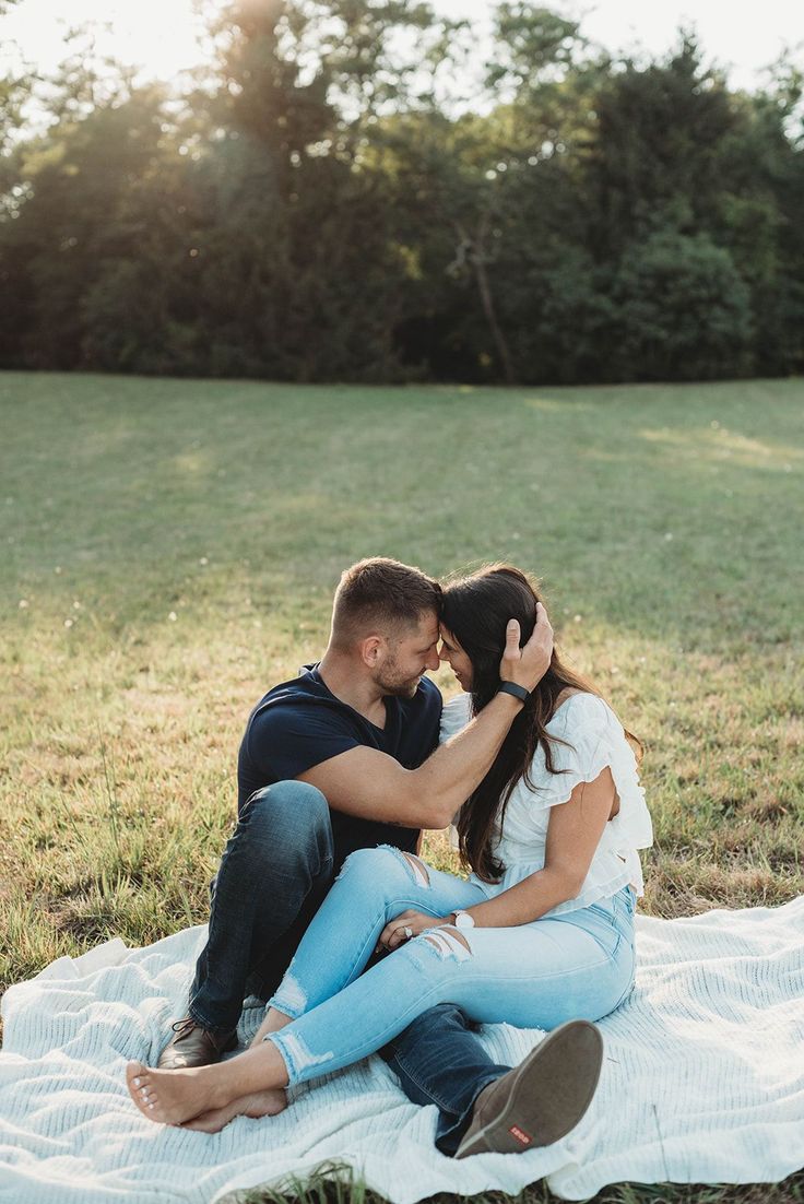 a man and woman sitting on top of a blanket in the grass with trees behind them