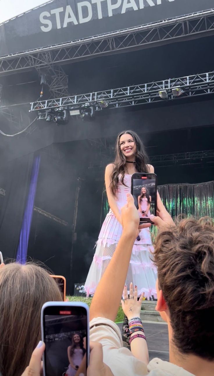 a woman in a white dress is holding up her cell phone while standing on stage