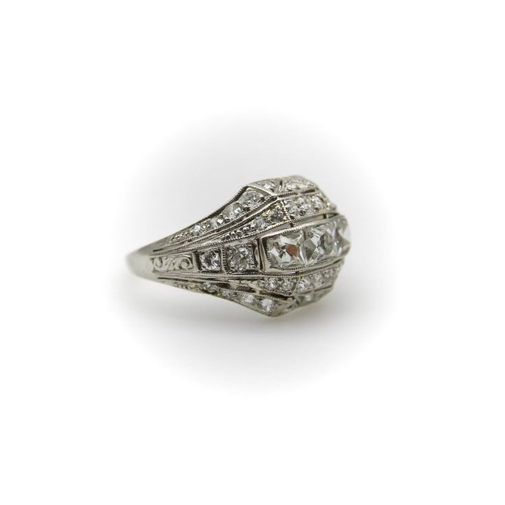 This platinum diamond ring is on the cusp of the Edwardian and Art Deco eras. The centerpiece of the ornate ring are three Peruzzi cut bezel set diamonds —one is an elongated rectangular shape, while the diamonds on either side are truncated, cut specifically for this ring. The larger diamonds are surrounded by a selection of 40 single and Old Mine Cut diamonds that are bead set into the ring. The ring has layers divided by milgrain work that tie together the lace-like patterns of the Edwardian era and the geometric designs of the Art Deco era in alternating bands. There is an added detail of a hand engraved scrolling foliate on each side of the ring. The ring has open work on the bottom that lets in the light and also reveals the triangular bottom shape of the diamonds—a nice touch that h Luxury Rings With Diamond Accents And Rectangular Stone, Luxury Rings With Rectangular Diamond Accents, Luxury Wedding Rings With Rectangular Stone, Luxury Rectangular Stone Wedding Ring, Formal Art Deco Square Cut Jewelry, Art Deco Platinum Diamond Ring With Rose Cut Diamonds, Art Deco Rings With Rectangular Stone For Formal Occasions, Heirloom Platinum Diamond Ring With Baguette Cut, Art Deco Baguette Cut Diamond Ring