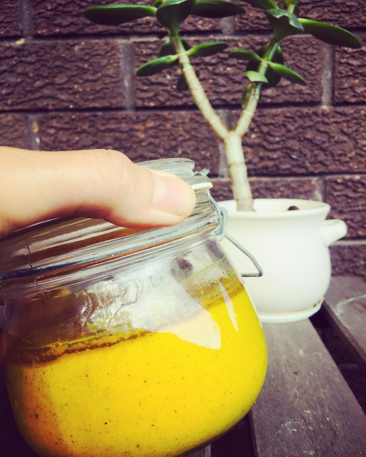 a hand is holding a jar full of yellow liquid next to a potted plant