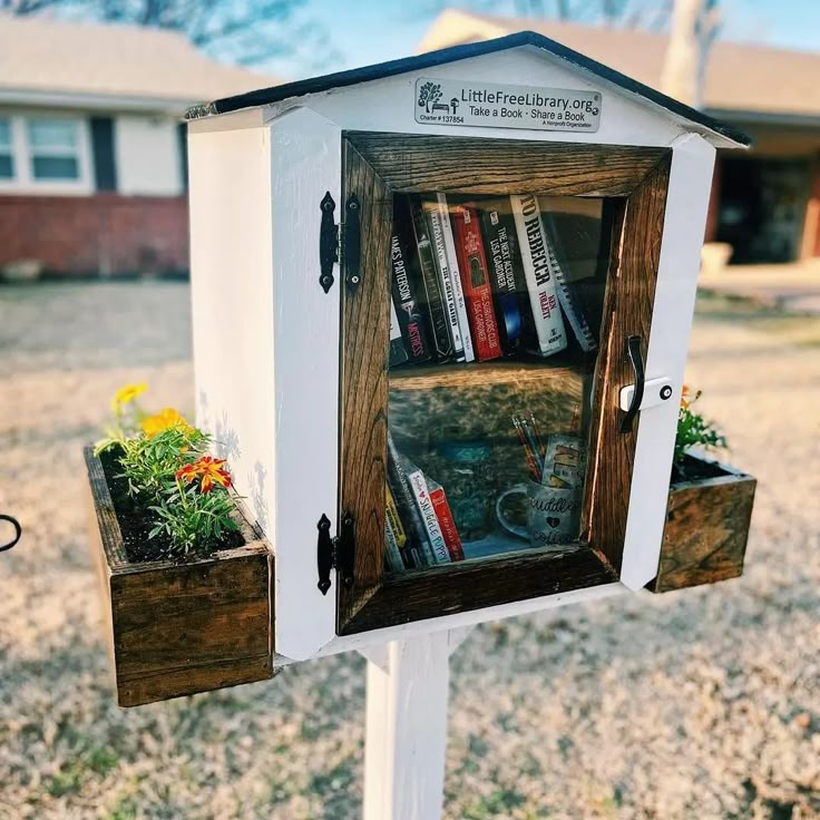 a wooden book shelf with books in it on top of a white post next to a yard