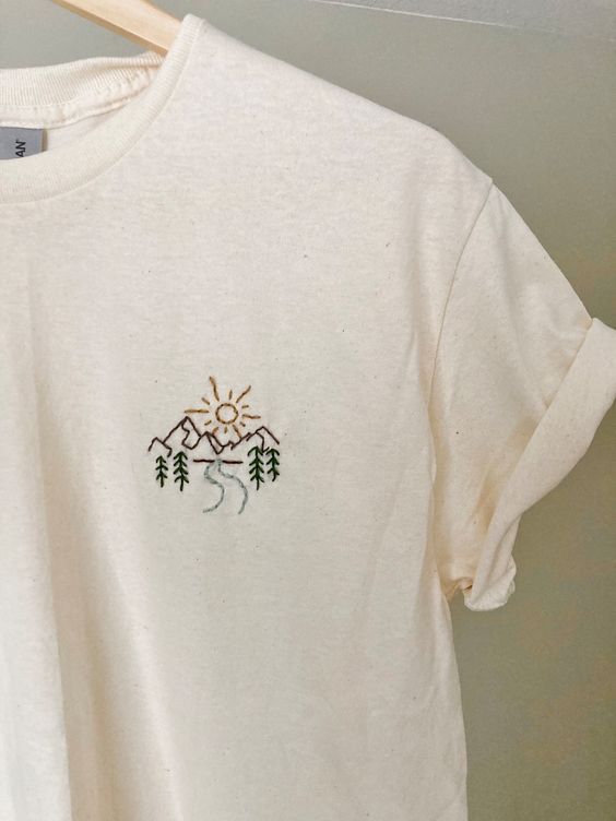 a white t - shirt with trees and mountains embroidered on the front, hanging from a wooden hanger