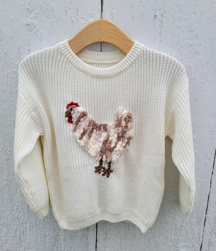 a white sweater with a red and brown poodle embroidered on the front, hanging from a wooden hanger