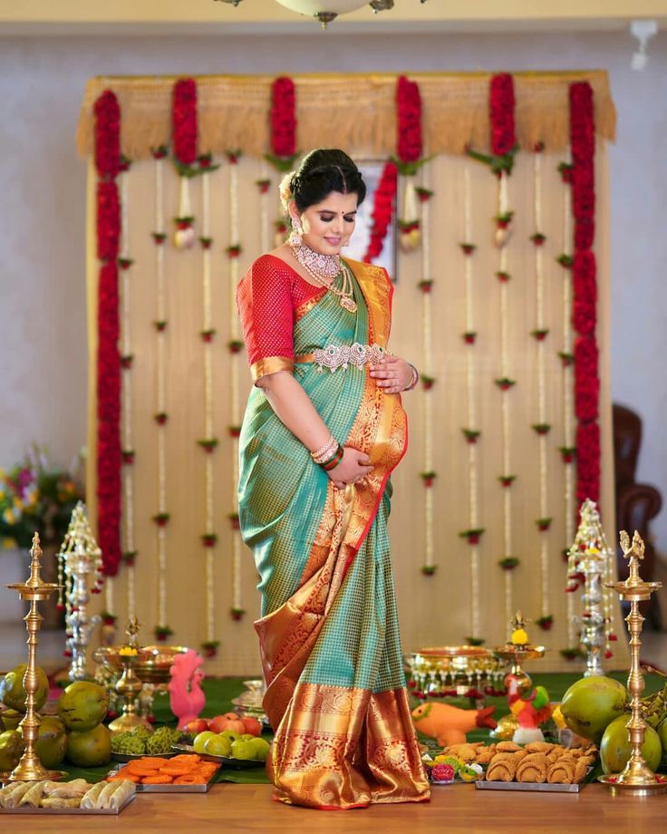 a woman in a green and gold saree standing on a stage with fruit around her