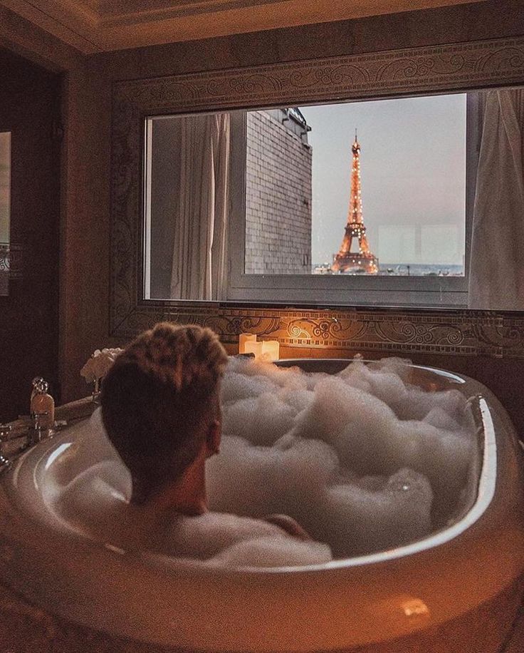 a man is sitting in the bathtub looking at the eiffel tower outside