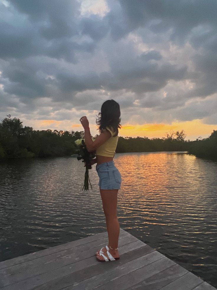 a woman standing on top of a wooden pier next to a body of water holding a bouquet of flowers