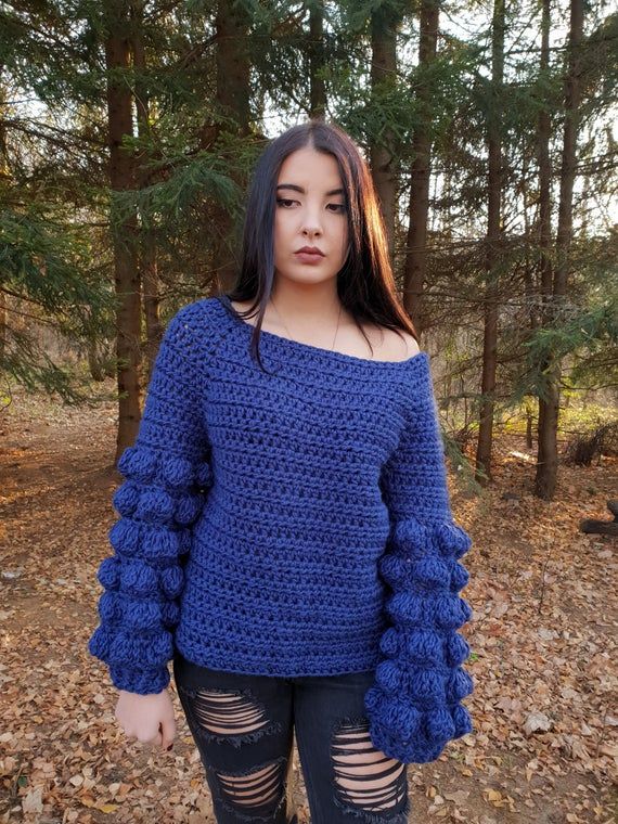 a woman standing in the woods wearing ripped jeans and a blue crochet sweater