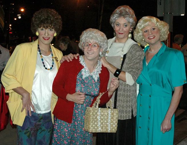 three women are posing for the camera with one woman holding a purse and two others wearing wigs