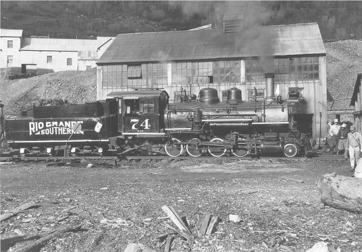 an old black and white photo of people standing near a train on the tracks in front of a building