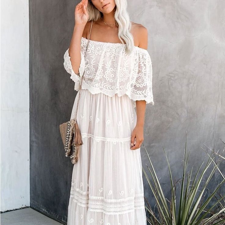 a woman standing in front of a wall wearing a white dress with off the shoulder sleeves