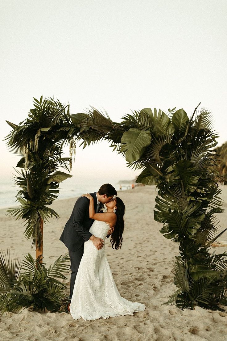 a bride and groom kissing under an arch made out of palm trees on the beach