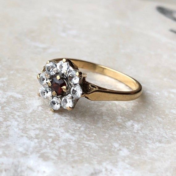 A vintage 9 carat gold paste (stones that mimic diamonds and rubies) ring. This beautiful piece holds a gorgeous red stone, framed by white stones, creating a floral cluster. This piece has simple shoulders, leading to a simple band. A lovely classic shape that is timeless!CONDITION: Wear consistent with age and use. Please see photos for more detail.ASSAYED IN LONDON, MADE IN CIRCA.1976APPROX. SETTING DIAMETER: 10mmAPPROX. SETTING HEIGHT: 6mmAPPROX. BAND WIDTH: 1.5mmRING SIZE: UK: N | US: 6 3/4