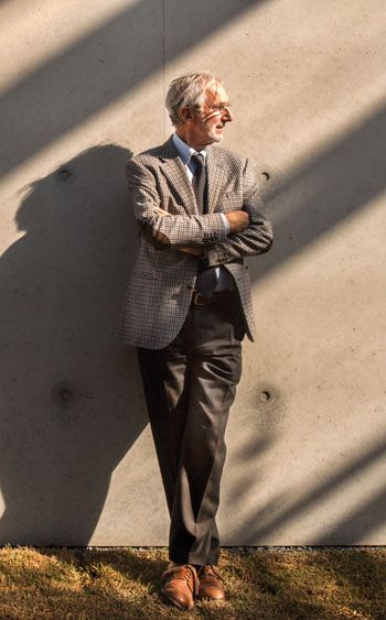 an older man leaning against a wall with his arms crossed, wearing a suit and tie