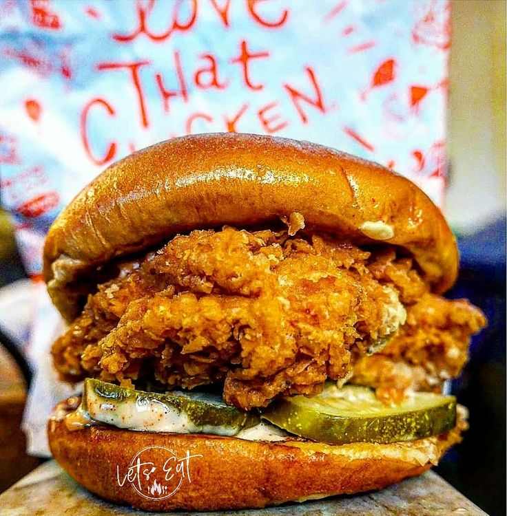 a fried chicken sandwich with pickles and cheese on a wooden cutting board next to a sign