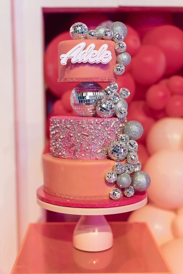 a three tiered cake with pink and silver decorations on the top, surrounded by balloons