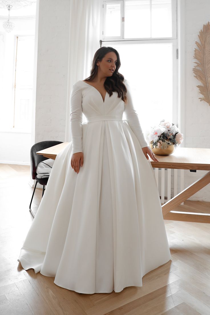 a woman standing in front of a window wearing a white wedding dress with long sleeves