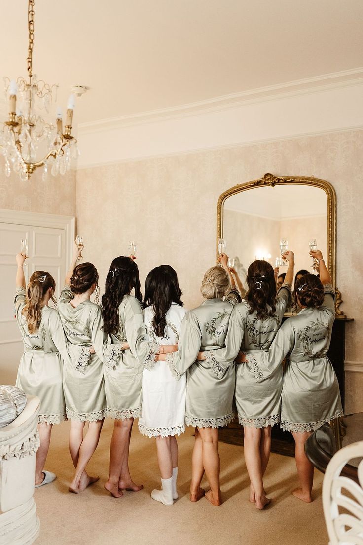 Bride wears white silk short dressing gown alongside her bridesmaids who wear matching dressing gowns in sage green Bridal Party Robes Sage Green, Sage Green Wedding Getting Ready, Brides With Bridesmaids, Bridesmaids Getting Ready Photos Robes, Wedding Dressing Gowns, Matching Robes For Bridesmaids, Sage Green Bridesmaid Getting Ready, Sage Robes Bridesmaids, Sage Green Wedding Robes