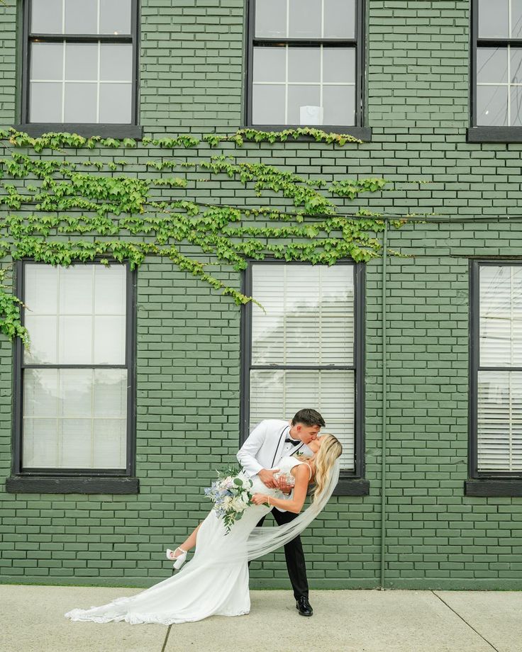 a bride and groom kissing in front of a green brick building