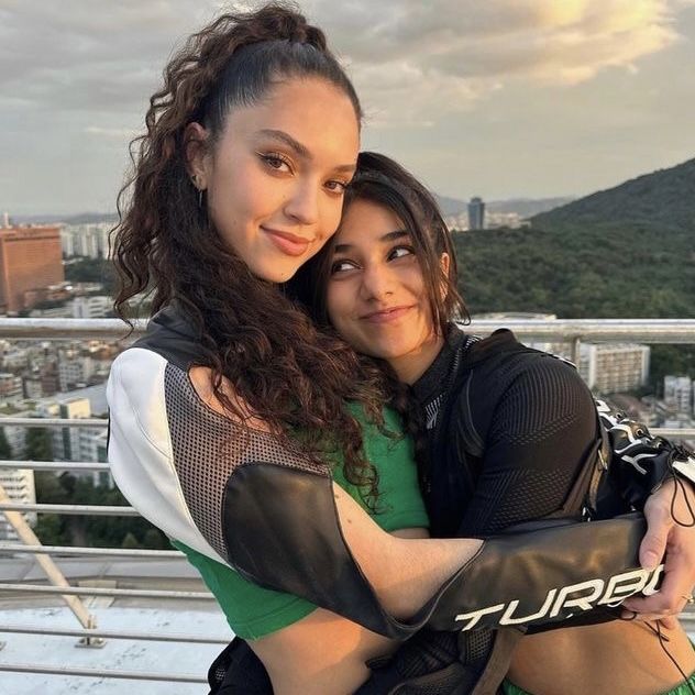 two young women hugging each other on top of a building with mountains in the background