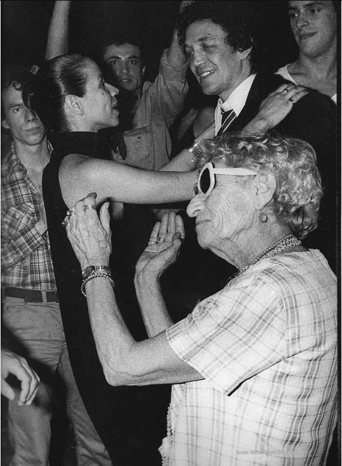 an older woman is dancing with her arm around the younger lady's shoulders while other people look on