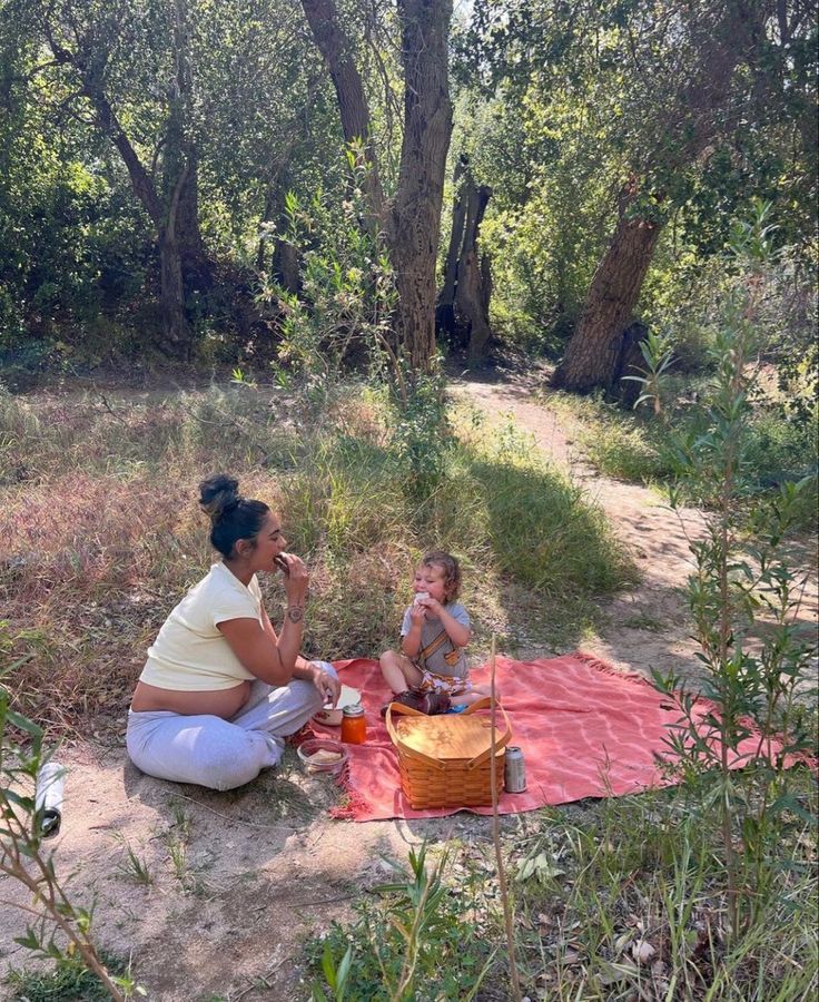 a woman and child are sitting on a blanket in the woods, eating food from a basket