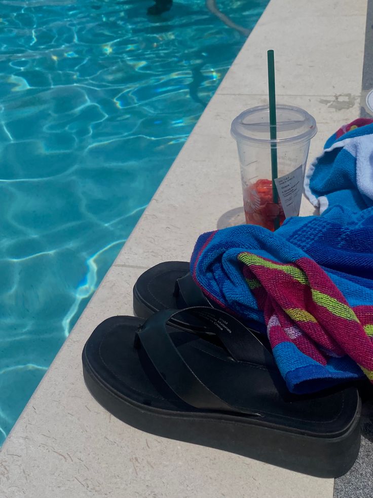 a pair of flip flops sitting next to a pool with a towel and drink