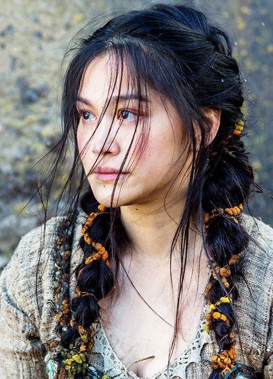 a woman with long black hair and braids on her head standing in front of a rock wall