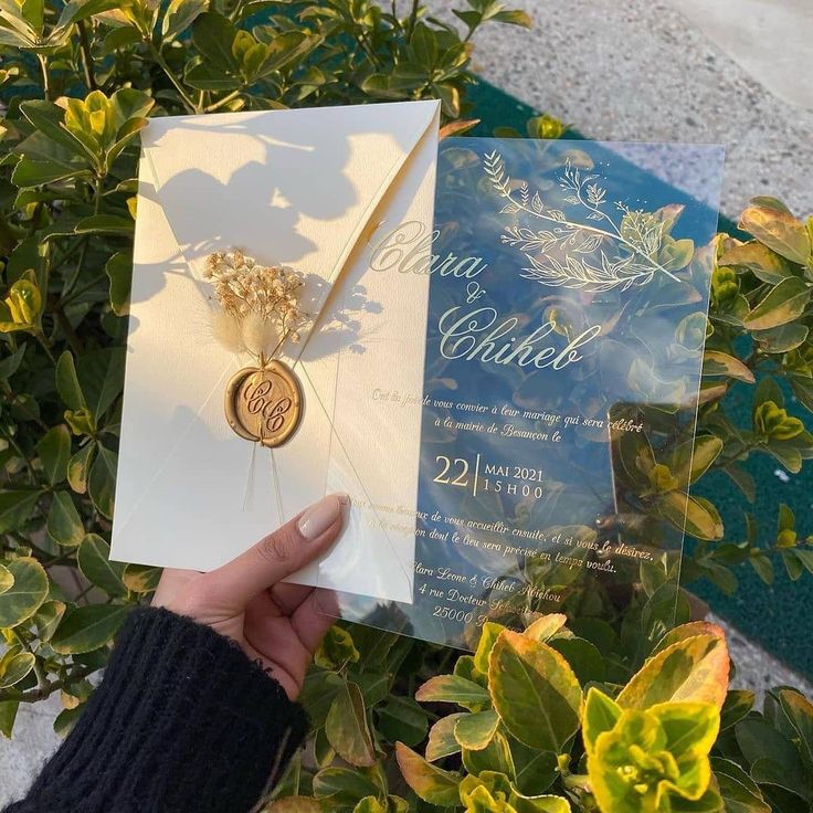 a person holding up a wedding card in front of some bushes and plants, with a gold brooch on it