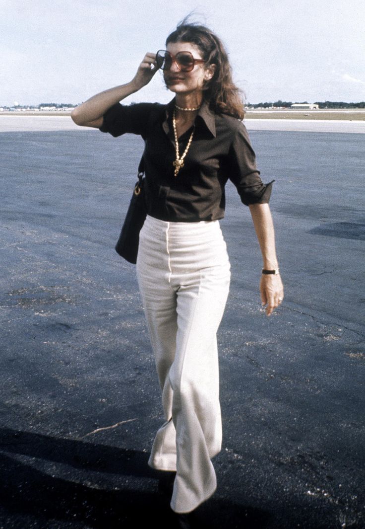 a woman walking across a parking lot while talking on a cell phone and wearing white pants