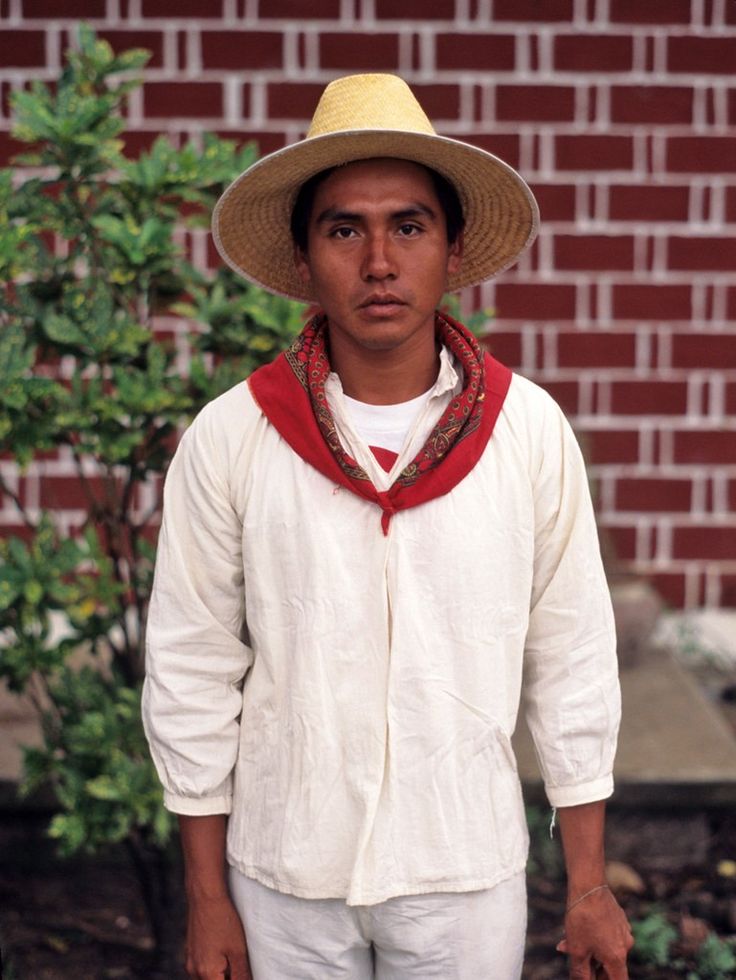 a man wearing a hat and white pants standing in front of a brick wall with a red scarf around his neck