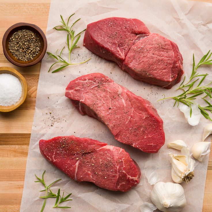 two raw steaks on parchment paper next to garlic and seasoning