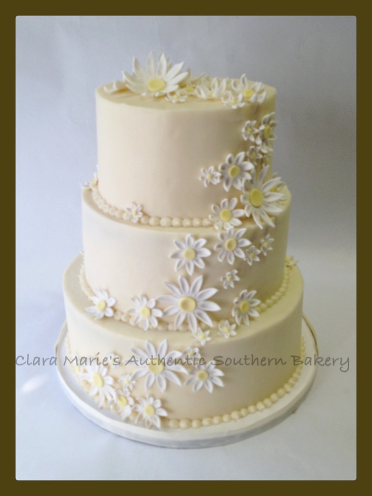 a three tiered cake with daisies and pearls on the top is decorated in white frosting