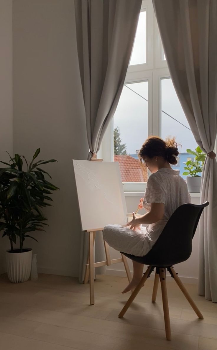 a woman sitting at a easel in front of a window