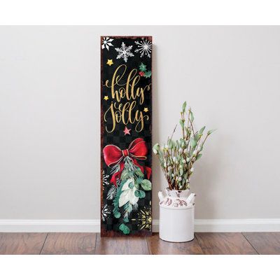 a christmas sign next to a potted plant on a wooden floor in front of a white wall