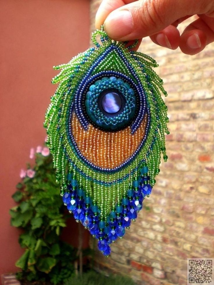 a hand holding a beaded peacock brooch in front of a brick wall and potted plant