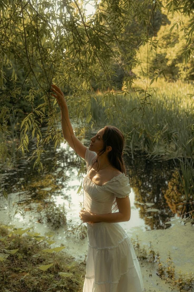 a woman in a white dress reaching up for leaves on a tree branch over water