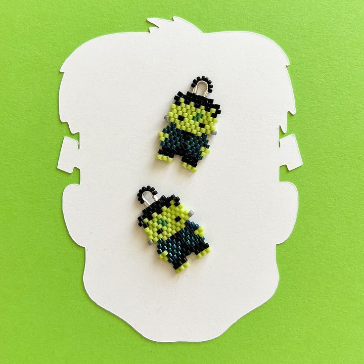 two small beaded earrings sitting on top of a white piece of paper next to a green background