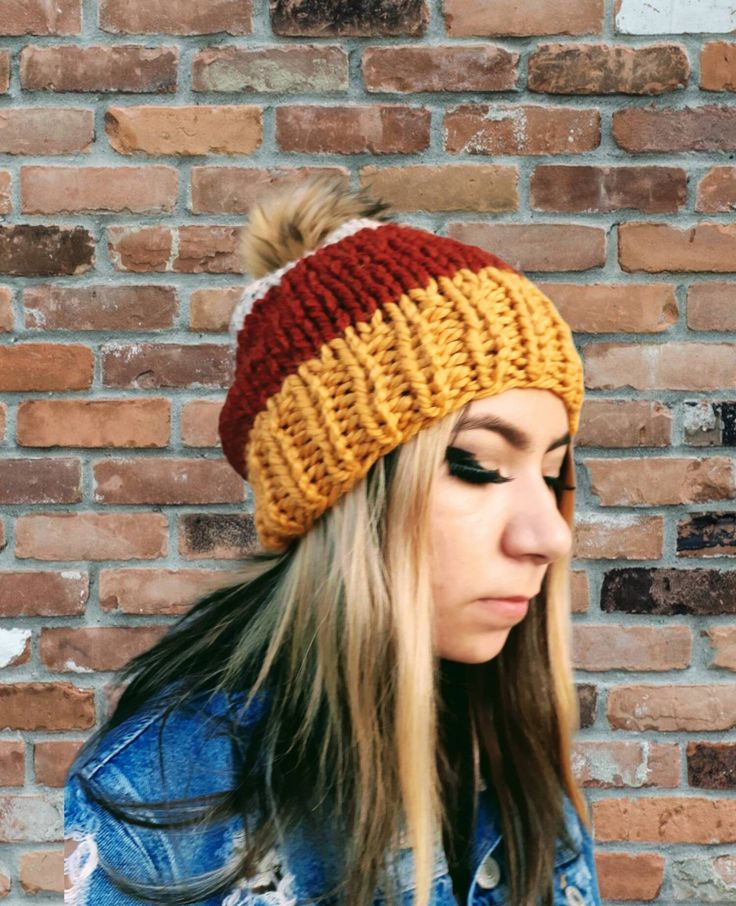 a woman wearing a knitted beanie with a pom in front of a brick wall