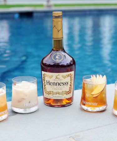 a bottle of hennesy next to four glasses filled with drinks near a swimming pool