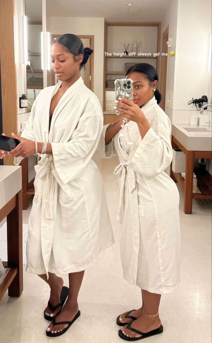 two women in white robes are looking at their cell phones while standing in the bathroom