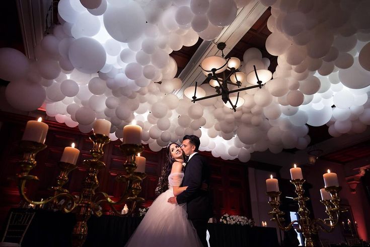 a bride and groom kissing under balloons in the ceiling at their wedding reception, with candles lit