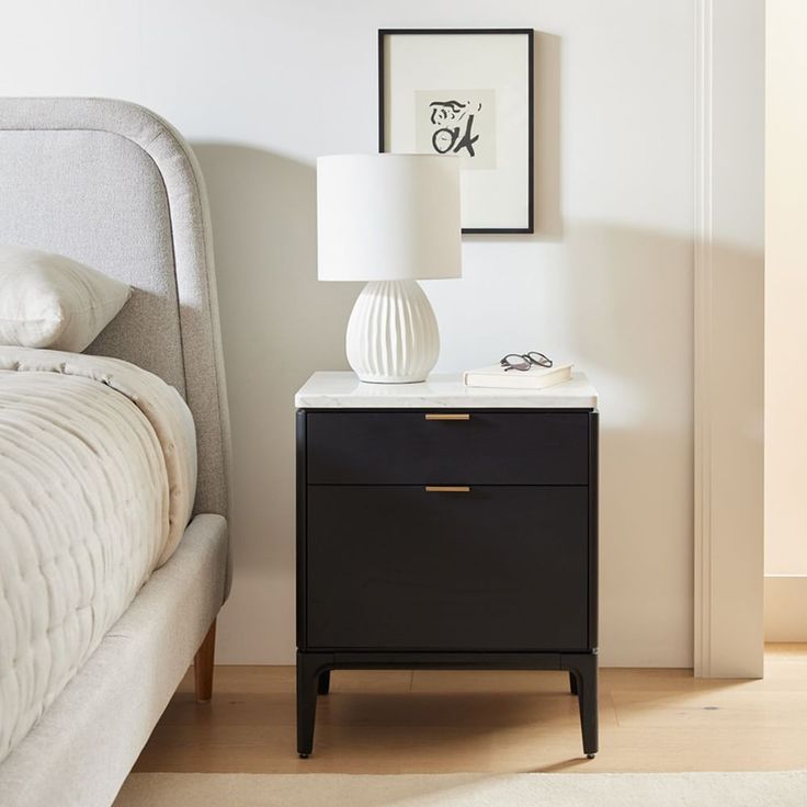 a nightstand with a lamp on top of it next to a bed and a painting