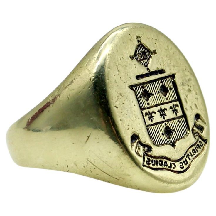 This 14k gold signet ring features a family crest of a shield topped with a radiating cross. The shield is divided into three sections, with vertical lines across the top and bottom sections, and a row of fleur-de-lis across the center. Above the shield is a cross like symbol that contains a tiny image that appears to be a book. Below the shield is a banner with the Latin phrase "Spiritus Gladius” which translates to "Sword of the Spirit.” A wonderful patina has settled into the signet’s engraving that adds depth to and delineates the images. The face of the ring measures 5/8” x 3/4” and the ring weighs 10.9 grams. Marked as 14k gold. Circa 1900, this makes a great unisex ring and could be sized up to fit a man—but we love it because the smaller ring size makes it perfect for a woman or so Classic Polished Signet Ring For Commemoration, Classic Engraved Signet Ring For Commemoration, Heirloom Engraved Signet Ring For Commemoration, Luxury Engraved Signet Ring For Commemoration, Engraved Oval Signet Ring For Commemoration, Classic Collectible Signet Ring With Coat Of Arms, Classic 14k Stamped Signet Ring For Commemoration, Heirloom Oval Signet Ring With Coat Of Arms, Classic Oval Signet Ring With Coat Of Arms