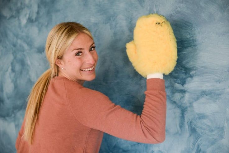 a woman holding a yellow sponge in her right hand and smiling at the camera while standing against a blue background