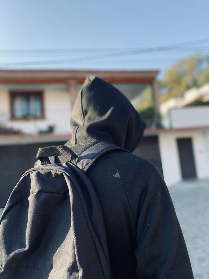 a person wearing a black hoodie with a back pack on their shoulder, walking towards a house