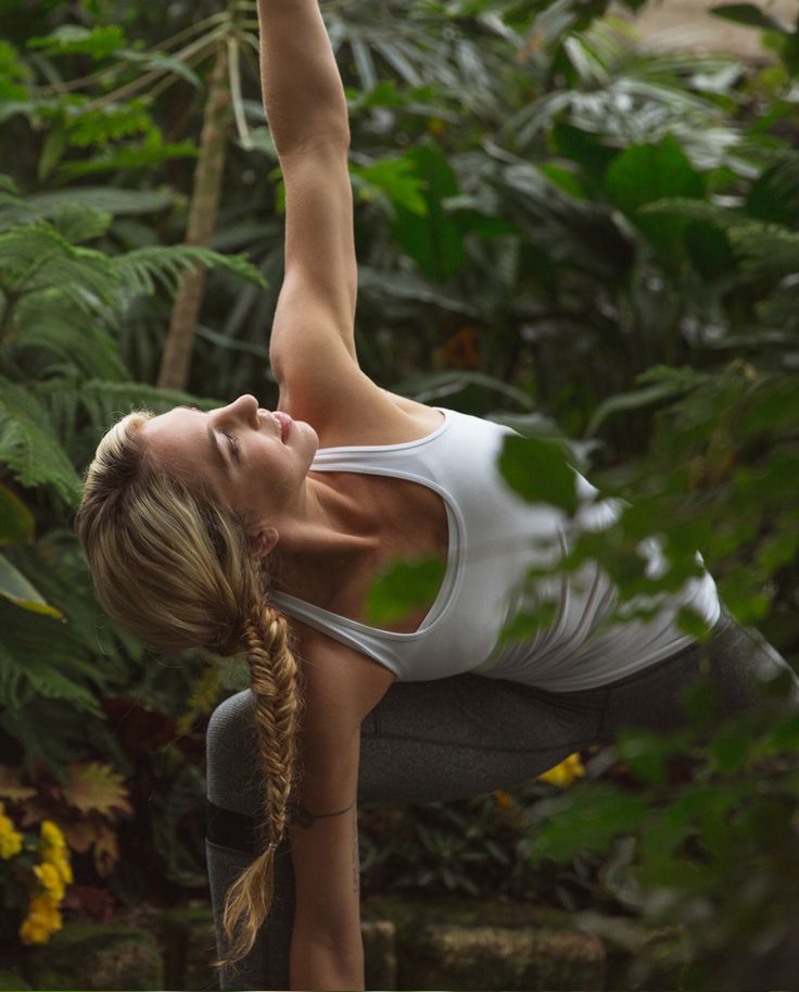 a woman is doing yoga in the middle of some plants and trees with her hands up