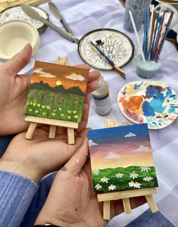 a person is holding two small paintings on wooden easels with paintbrushes in front of them