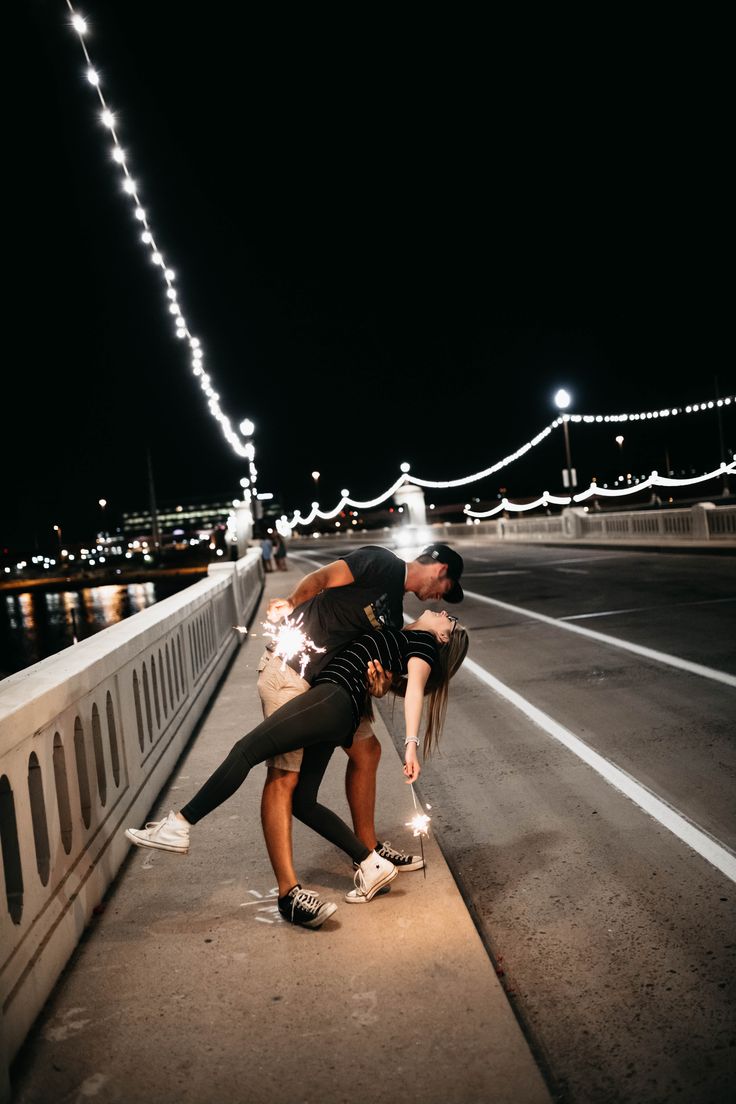 a man and woman kissing on the side of a bridge at night with lights in the background