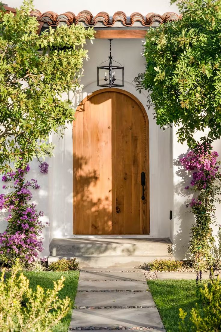 a wooden door in front of a white house with purple flowers on the side walk