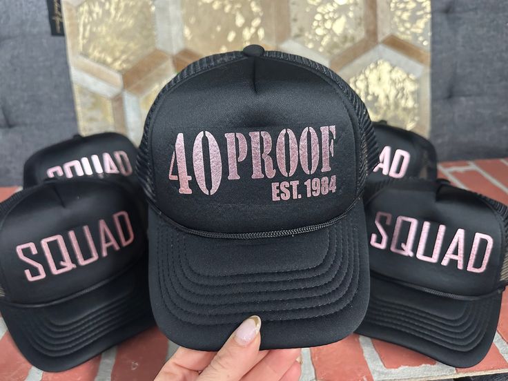 four black hats with pink letters and the words squadron est 1994 printed on them, sitting on a table